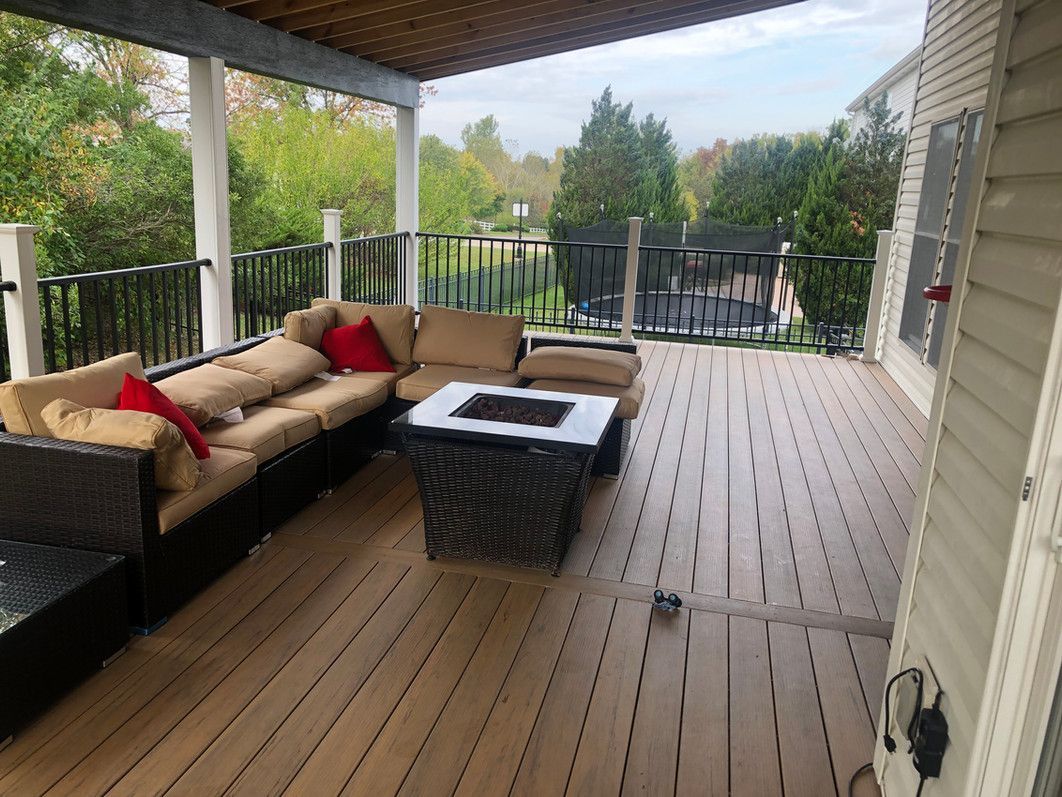 A covered wooden deck with outdoor furniture, including a sofa and fire pit. Trees and a fence are in the background.