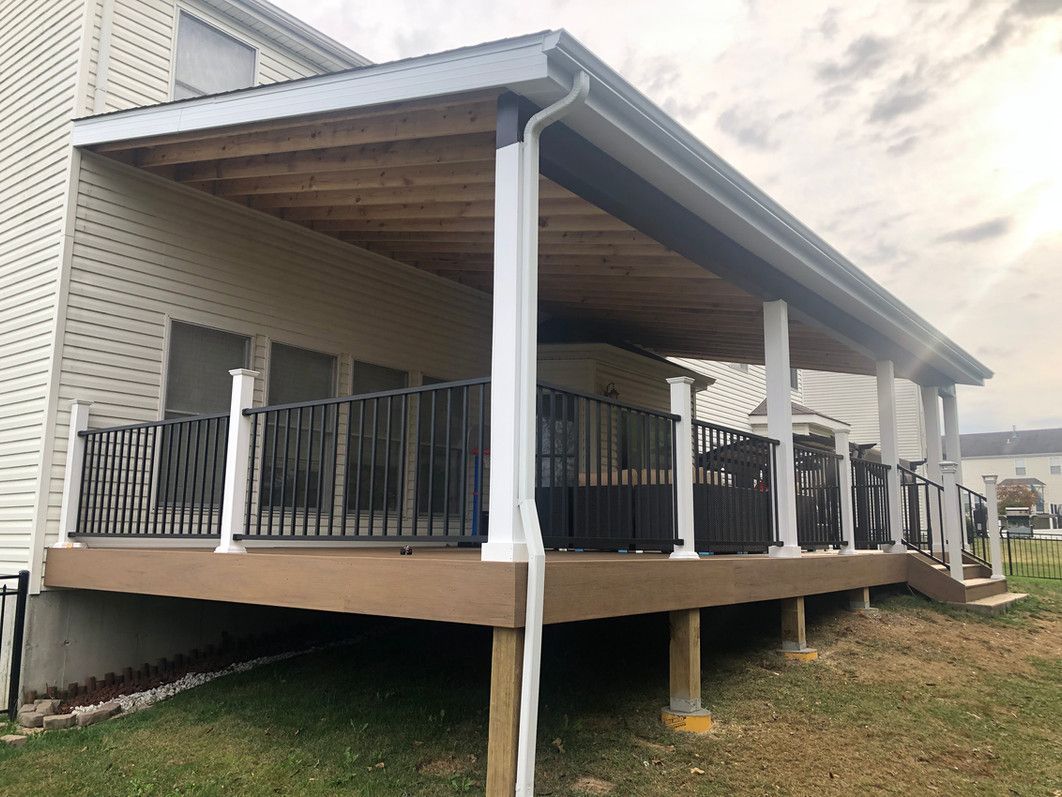 Covered deck with white columns, brown railing, and light-colored siding against a cloudy sky.