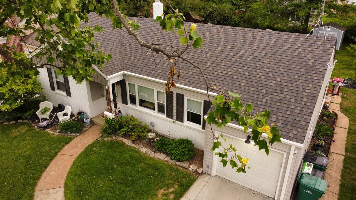 A one-story white house with a brown roof and a curved walkway; a tree partially obscures the roof.
