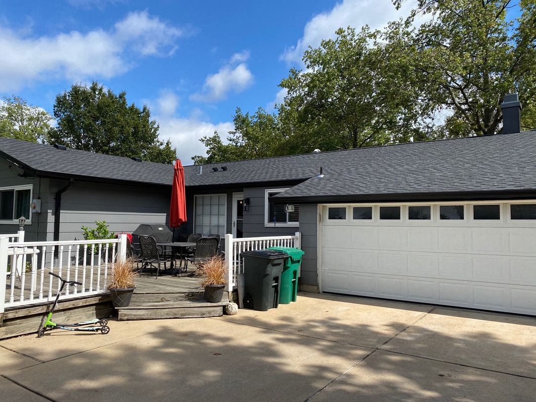Gray house with white garage door and deck, black roof, blue sky with some clouds.