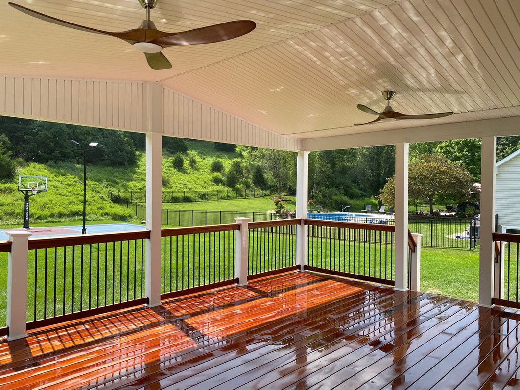 Covered porch with wood deck, ceiling fans, black railing, and view of a green landscape and pond.