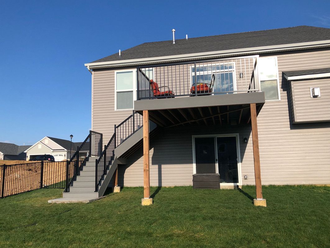Two-story house with a wooden deck and stairs; red chairs on deck, gray and brown features, grassy yard, and blue sky.