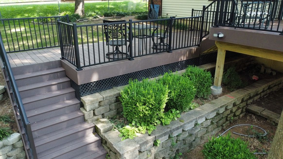 Outdoor deck with steps, black railing, brown boards, and small green bushes against a stone wall.