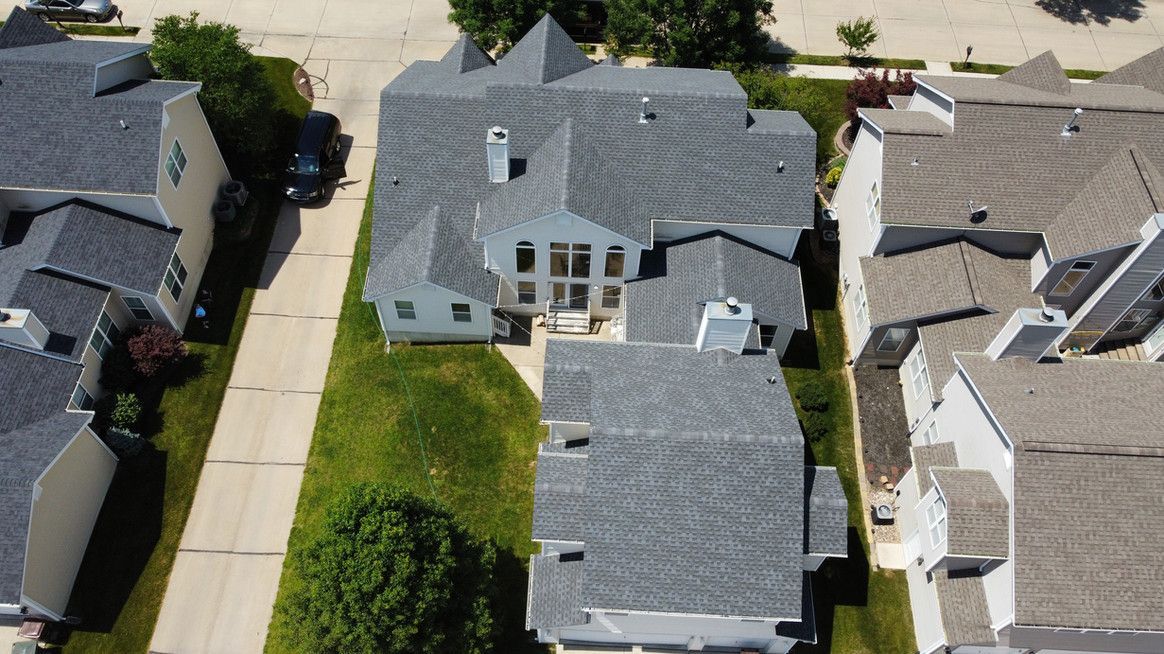 Aerial view of a gray-roofed house with lush green lawn, next to a paved road and other similar houses.