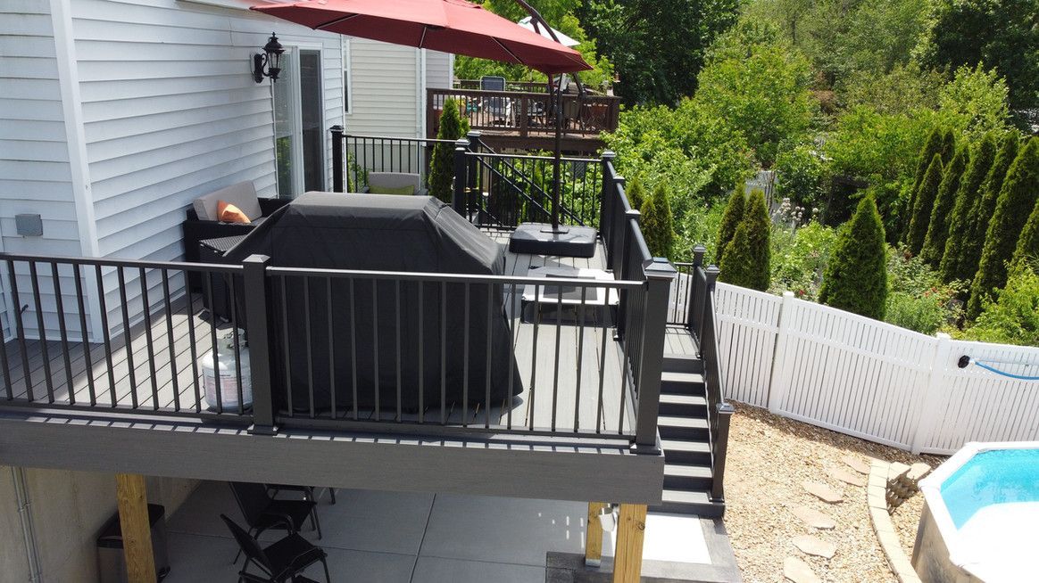 Deck with black railing, grill, umbrella, and steps leading down to a pool and greenery.