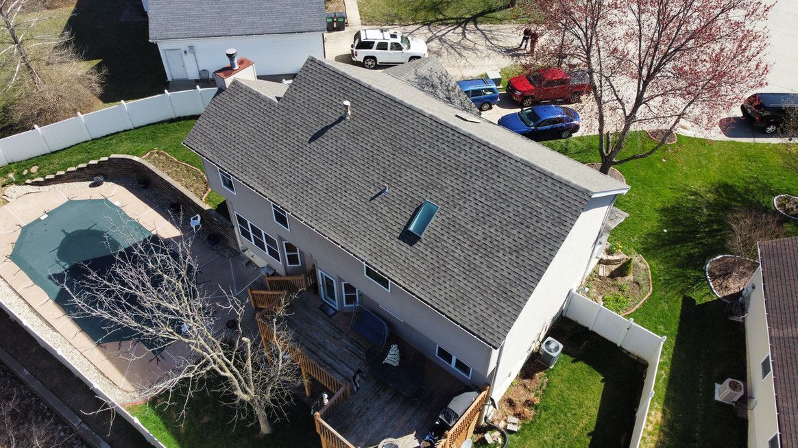 Aerial view of a gray-roofed house with a deck, swimming pool, and white fence in a green yard.