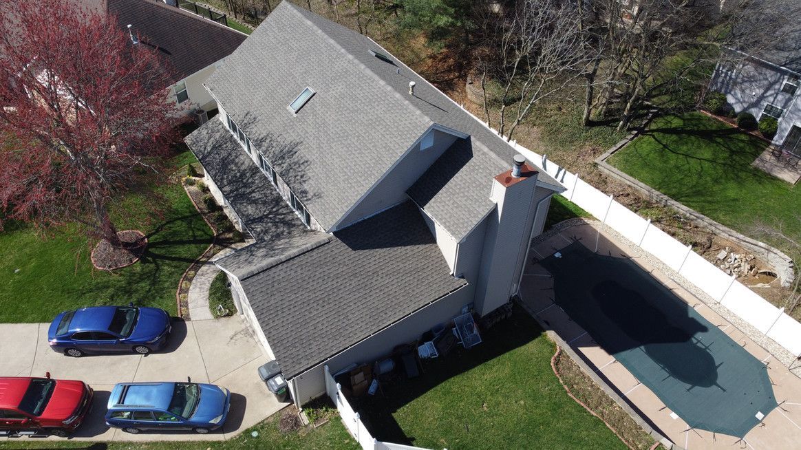 Aerial view of a house with a gray roof, pool covered in a tarp, cars parked in front, and green grass.