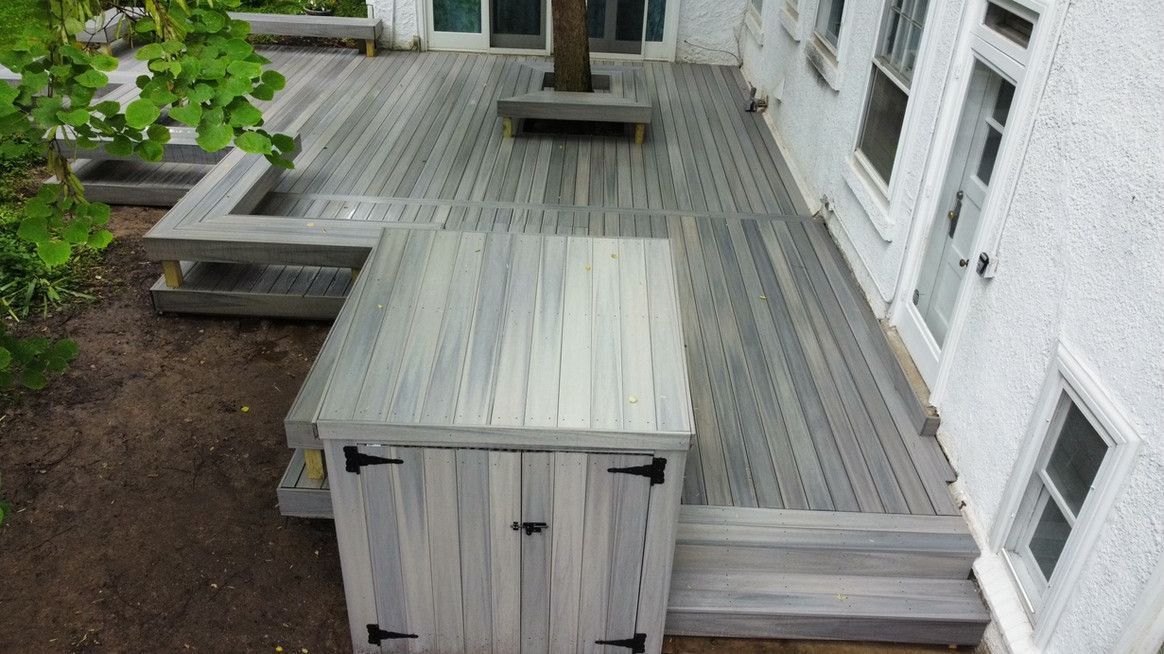 Wooden deck with built-in bench, surrounded by a white stucco building, a tree rises through the deck.