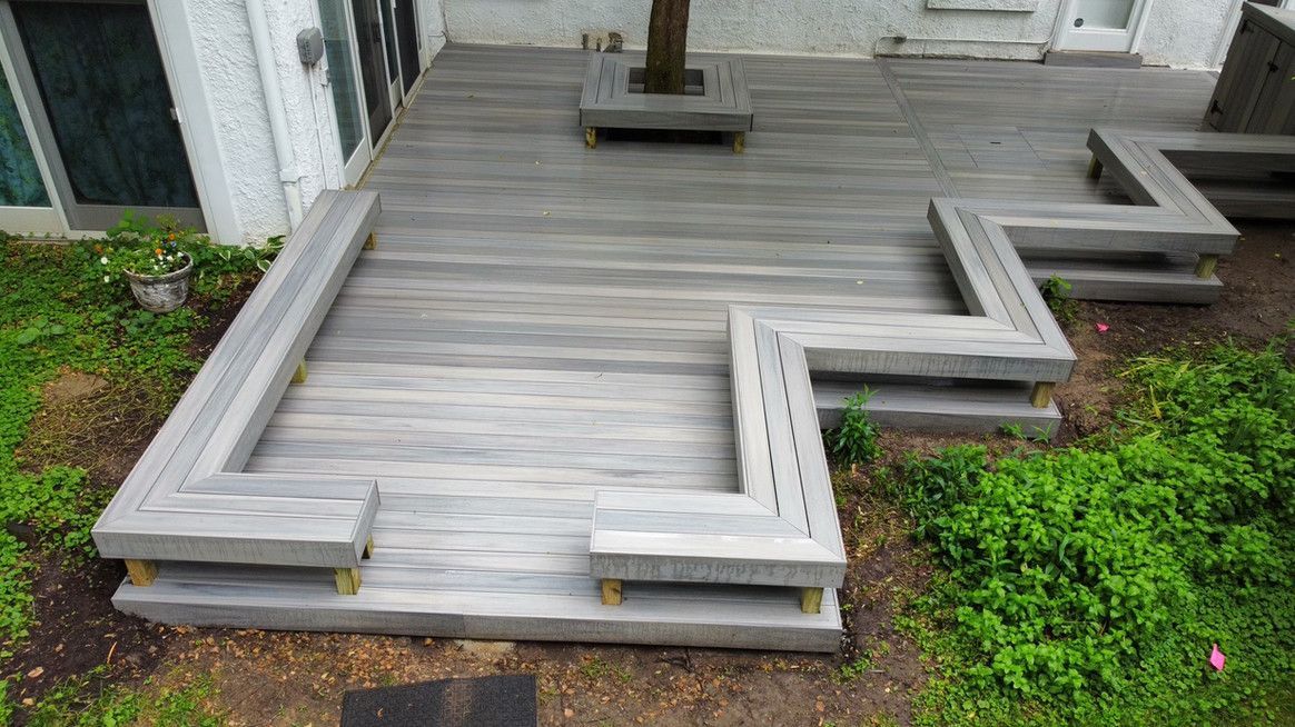 Wooden deck with built-in seating; gray planks, steps, and benches; near a building and greenery.