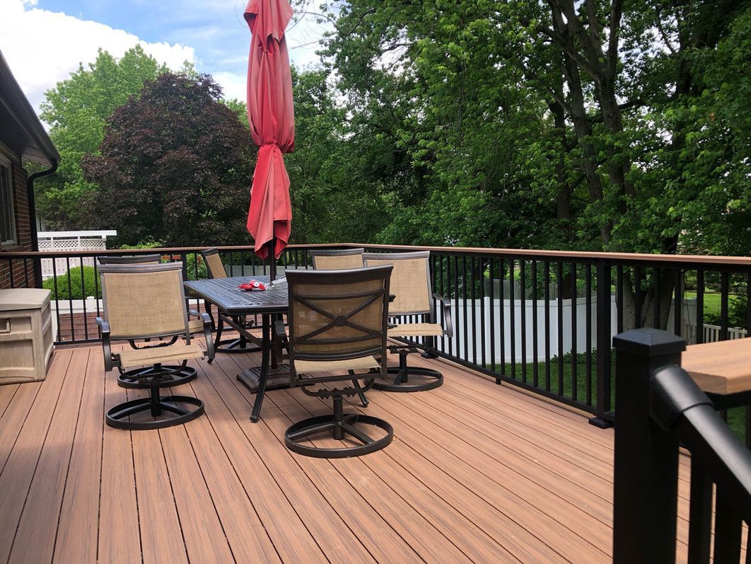 Wooden deck with outdoor dining set under a red umbrella, surrounded by black railing and trees.