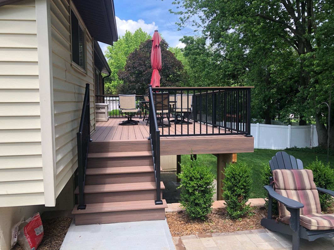 Backyard deck with stairs, composite decking, black railing, and red umbrella. Outdoor seating and green bushes.