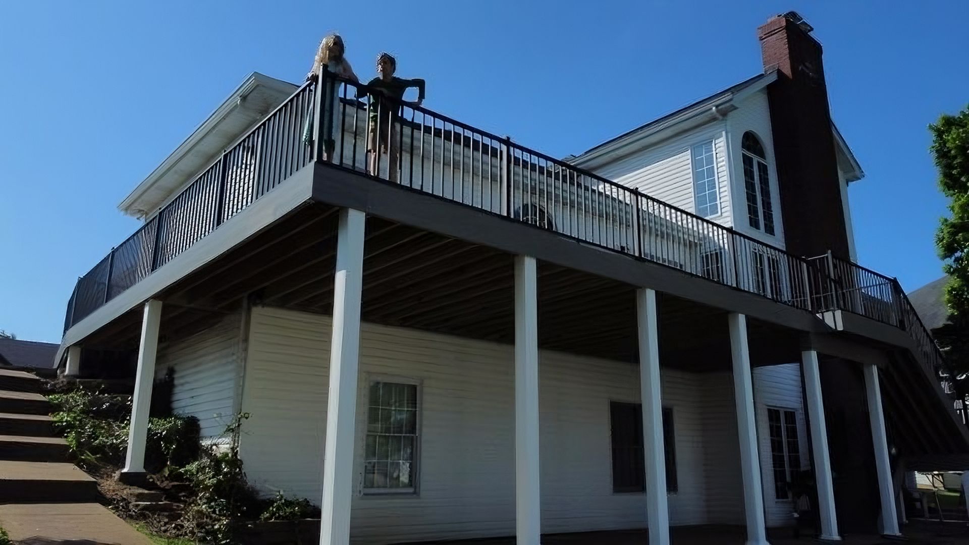 Two people on a large deck of a white house with a chimney; sunny day.