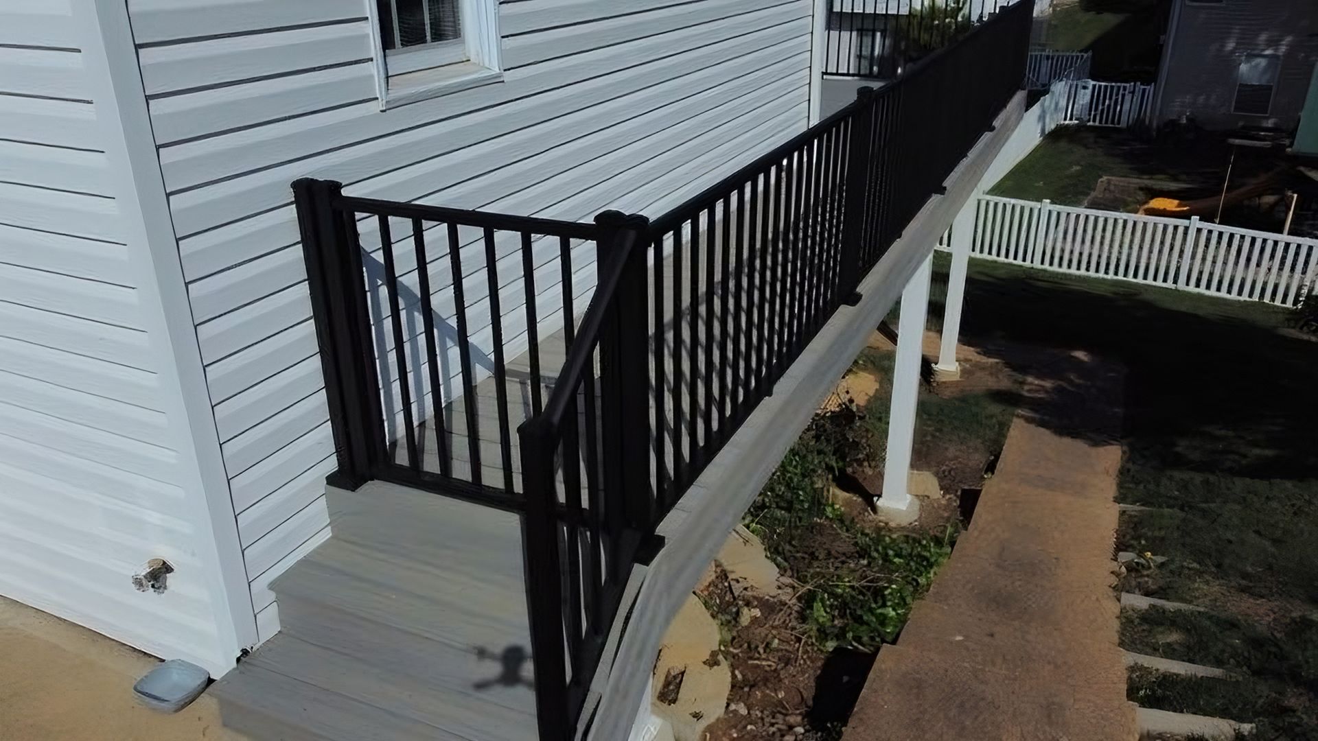 Black railing on a gray deck next to a house with white siding and a small yard.