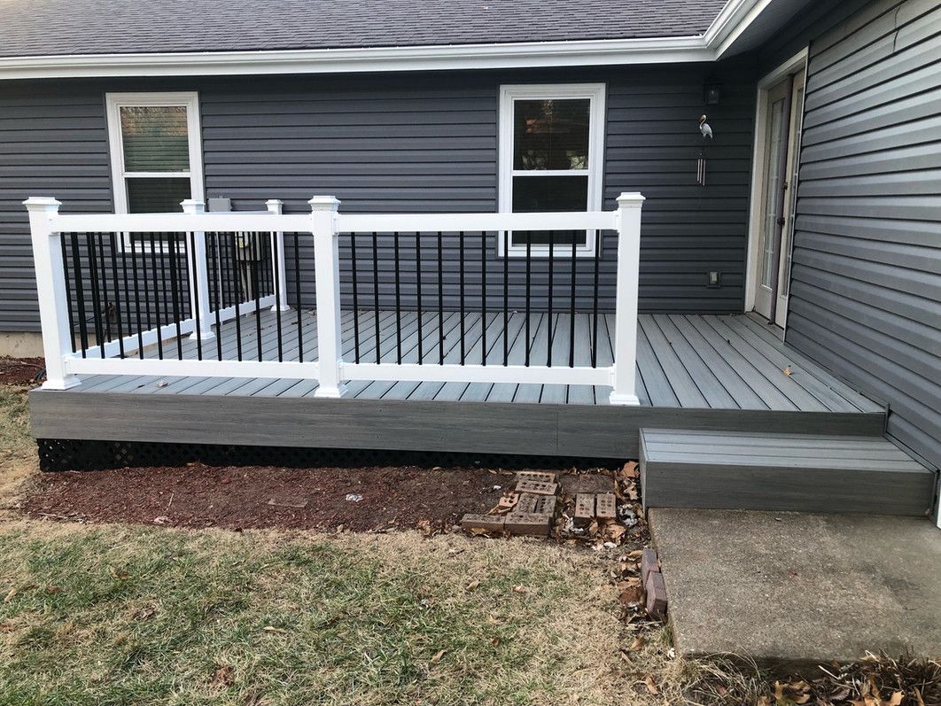 Gray deck with white railing, black spindles, and a step. Houses has gray siding and two windows.