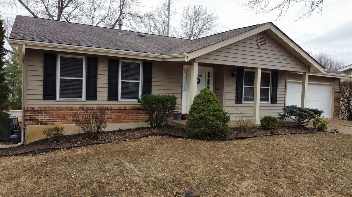 Tan ranch-style house with dark shutters, brick facade, and brown roof. Front yard with brown grass and landscaping.