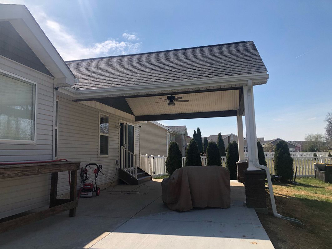 Covered patio attached to a light-colored house. Table under a brown cover. Green trees in the background.