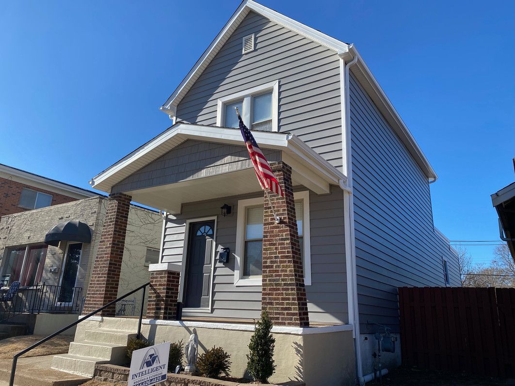Two-story gray house with an American flag. Brick pillars frame the porch. Sunny, blue sky.
