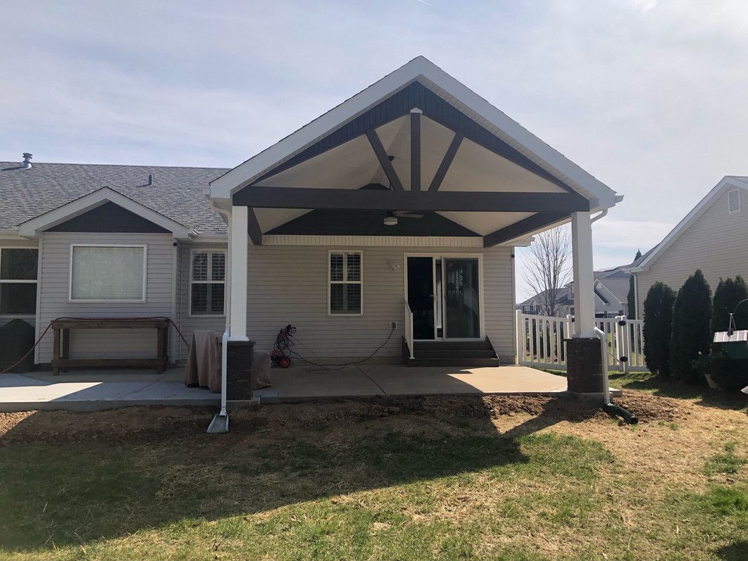 Backyard patio with a covered seating area. Light-colored siding, dark brown beams, gray roof, and green lawn.