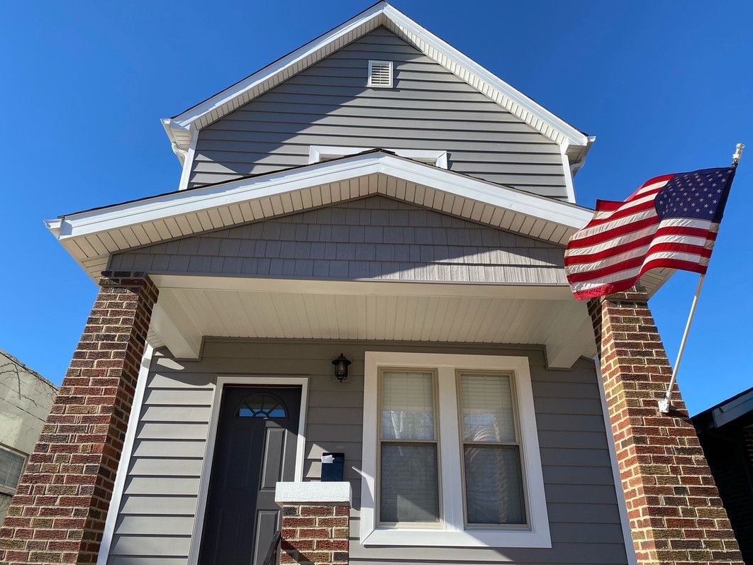 Two-story house with gray siding, brick columns, and an American flag on a sunny day.
