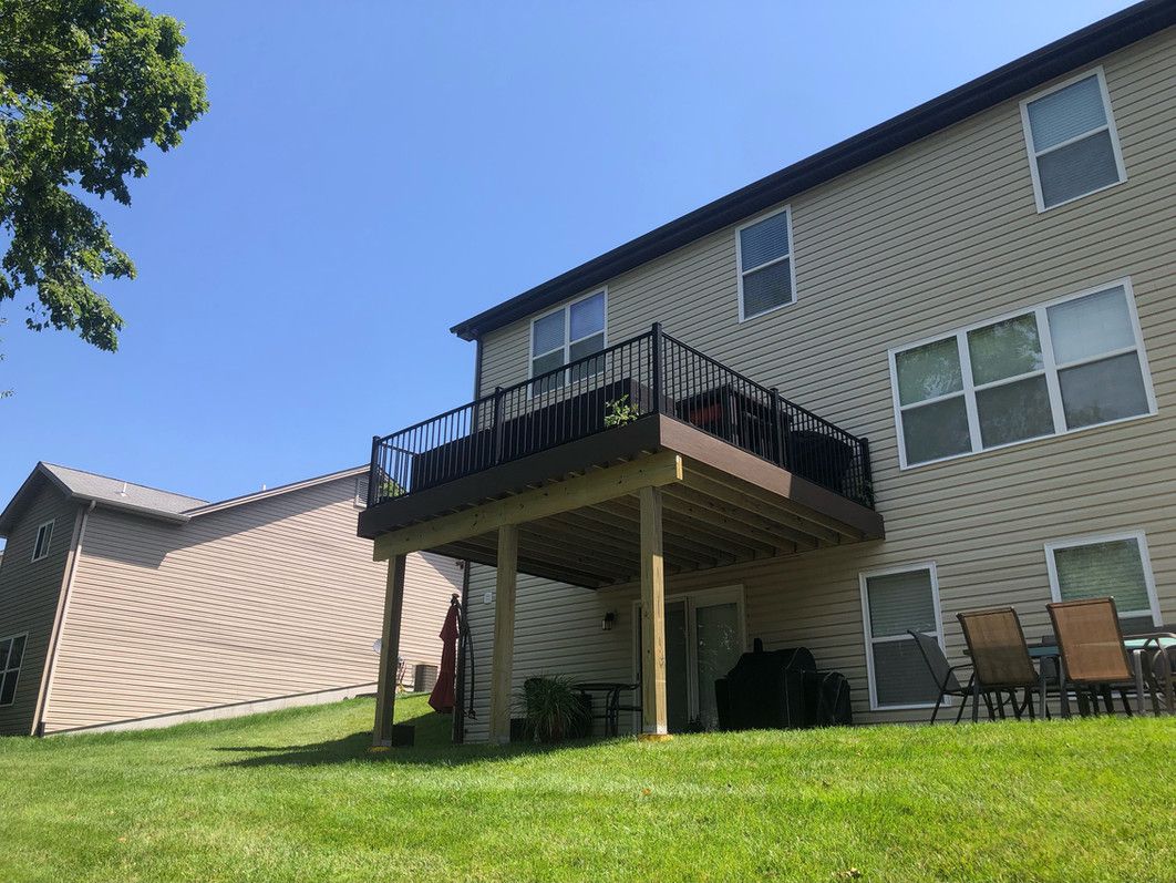 A two-story beige house with a brown deck, overlooking a grassy yard under a blue sky.