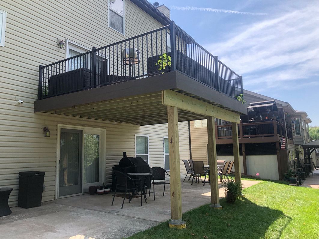 Two-story deck with black railing and brown boards, over a concrete patio and a grassy yard.