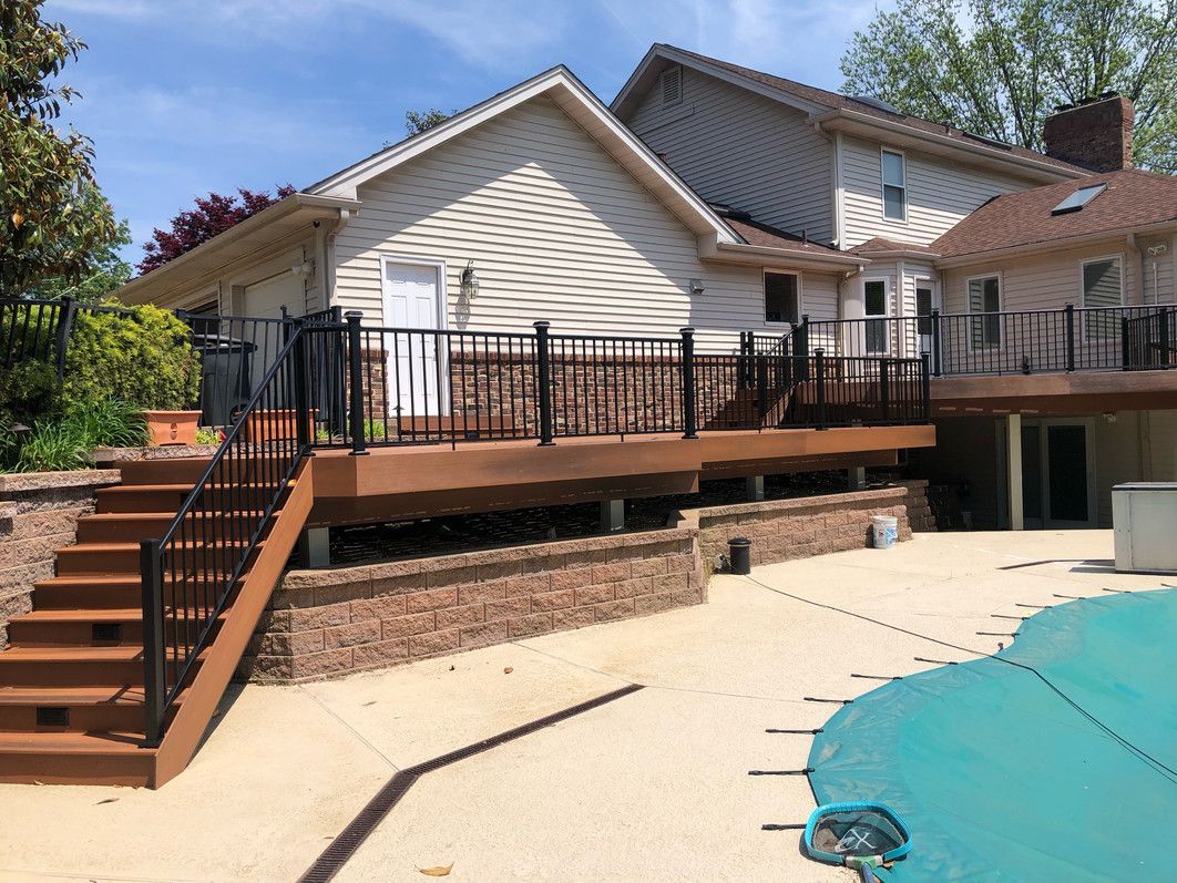 Deck and stairs of a house with black railings, tan deck, and pool partially visible on a sunny day.