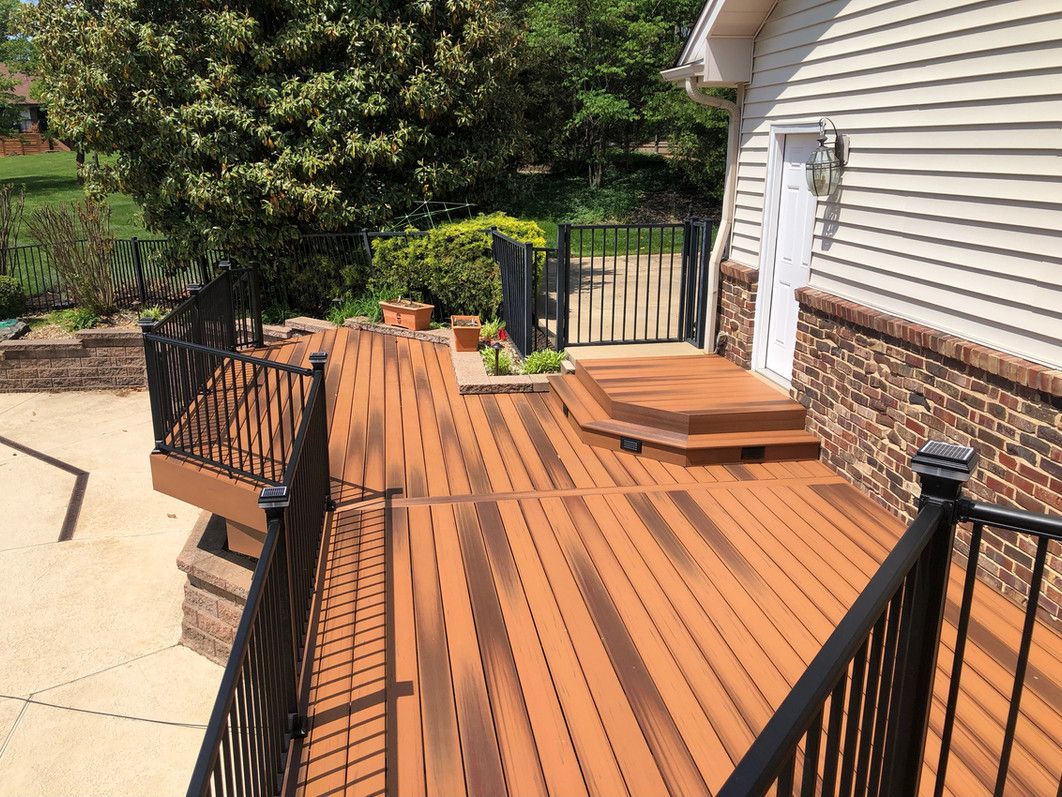 Brown wooden deck with black railings next to a brick-faced house, steps leading to a gate.