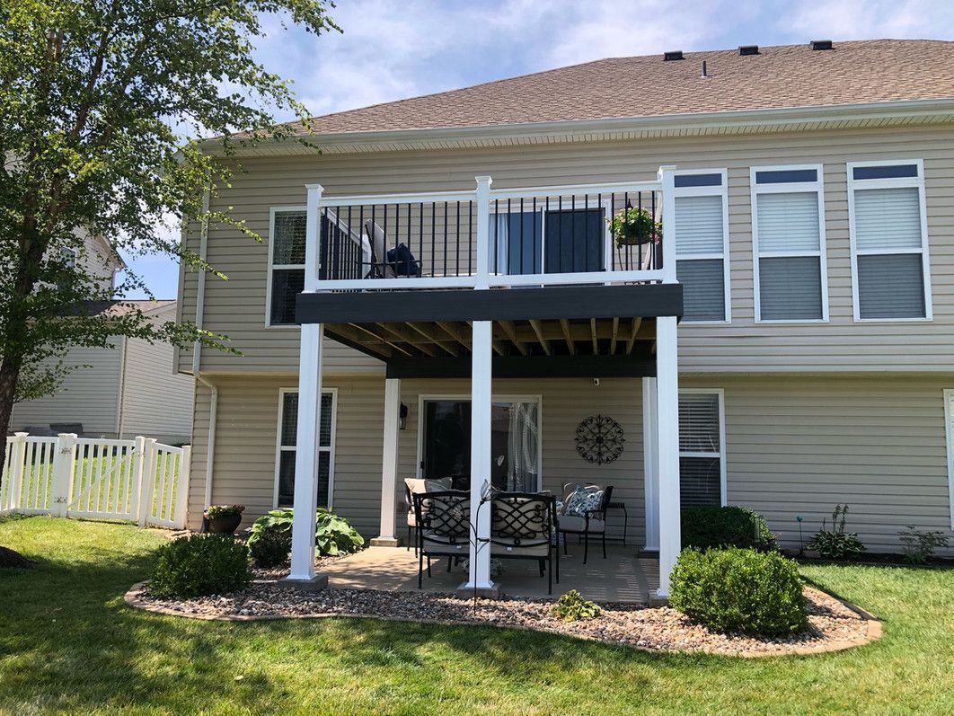 Two-story beige house with deck and patio. White columns support the upper deck. Patio furniture sits below.