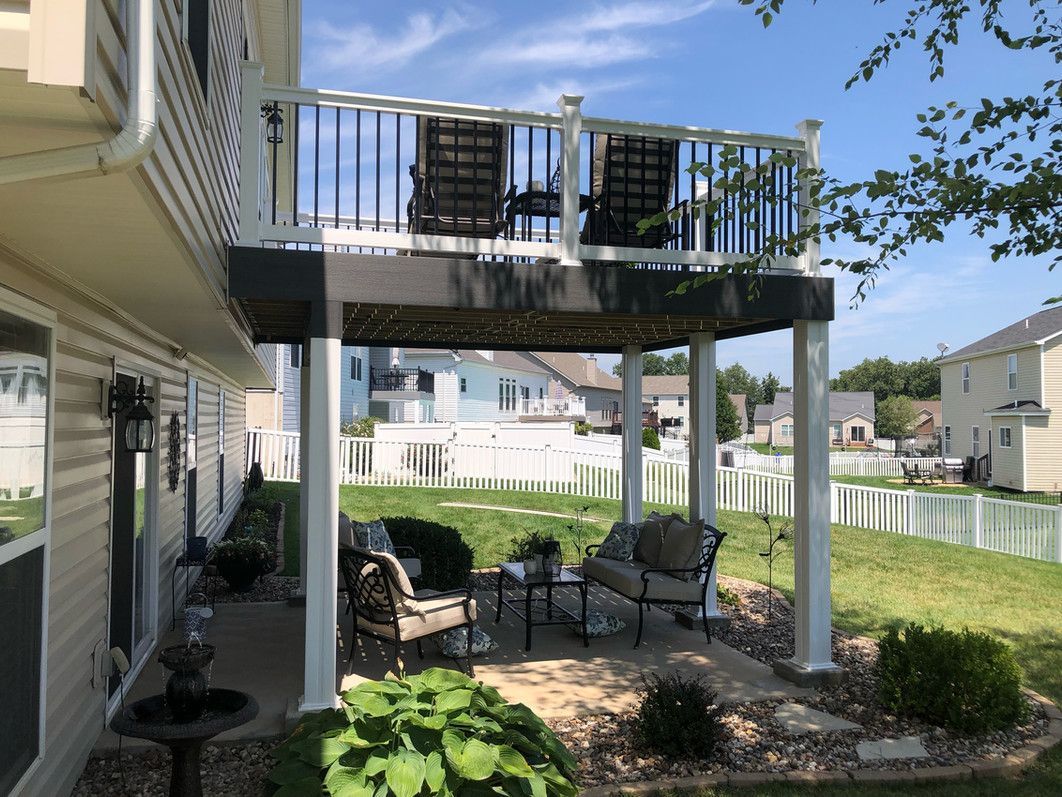 Two-level deck with seating. Upper level has chairs, lower level patio has furniture. White fencing, green grass. Sunny day.