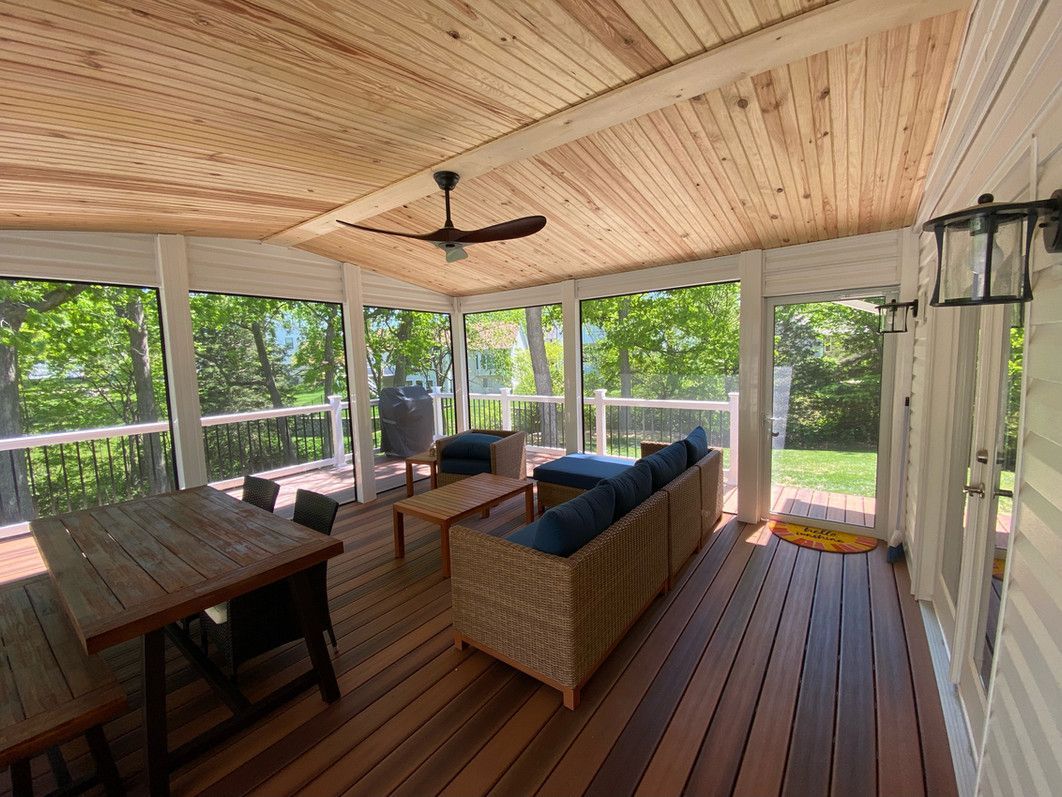 Screened-in porch with wood ceiling and floor, furnished with seating and dining table. Green trees visible through screens.