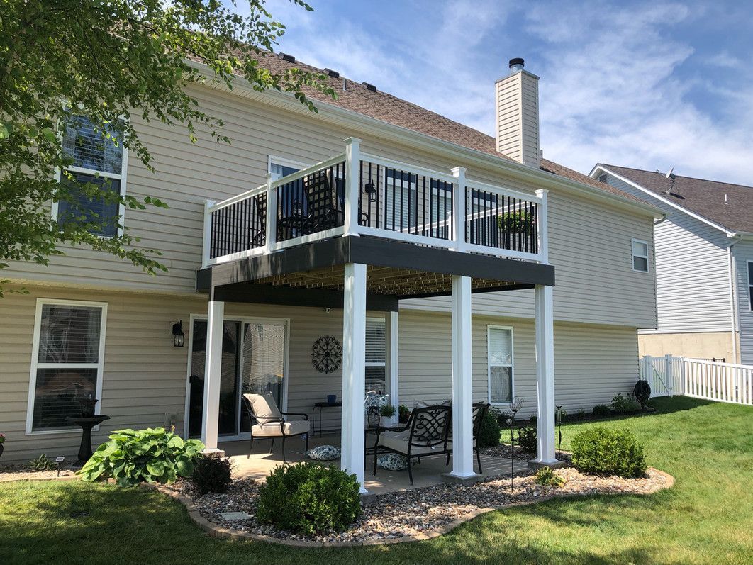 Two-story house with an elevated deck, black and white railing, and a patio below with seating.