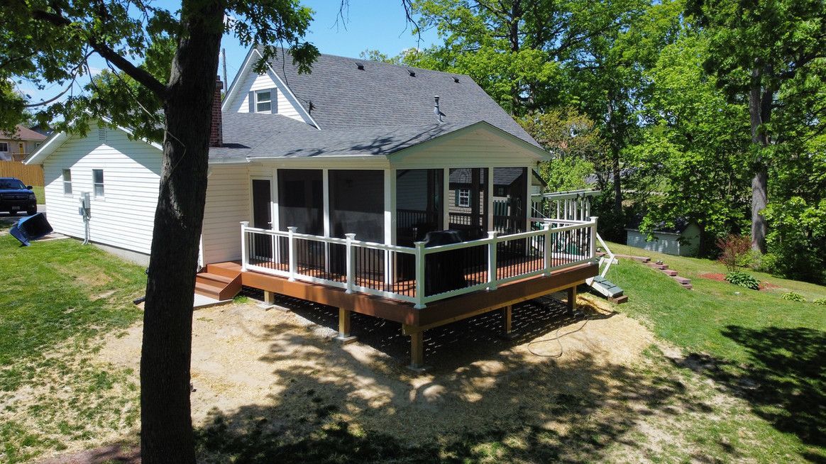 Cottage with screen porch and wooden deck surrounded by trees on a grassy hill.