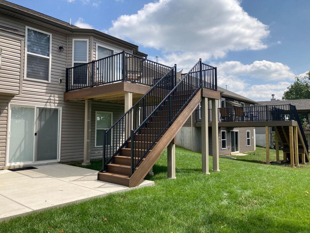 A deck with stairs attached to a two-story building.  Black railings, brown stairs, and a green lawn.