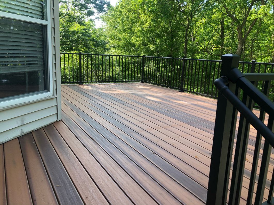 Wooden deck with black railings overlooking a treeline, next to a window with closed blinds.
