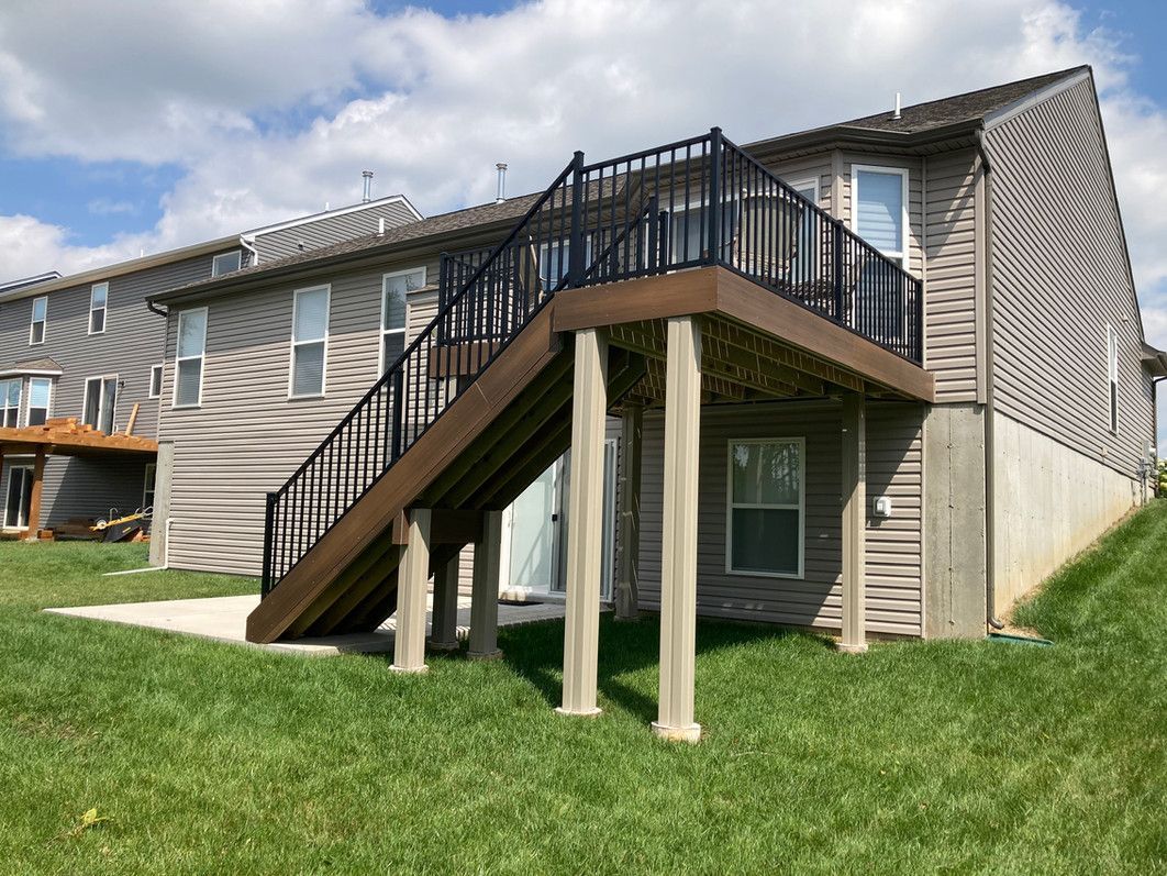 Brown deck with black railing and stairs attached to a two-story building on a grassy lawn.