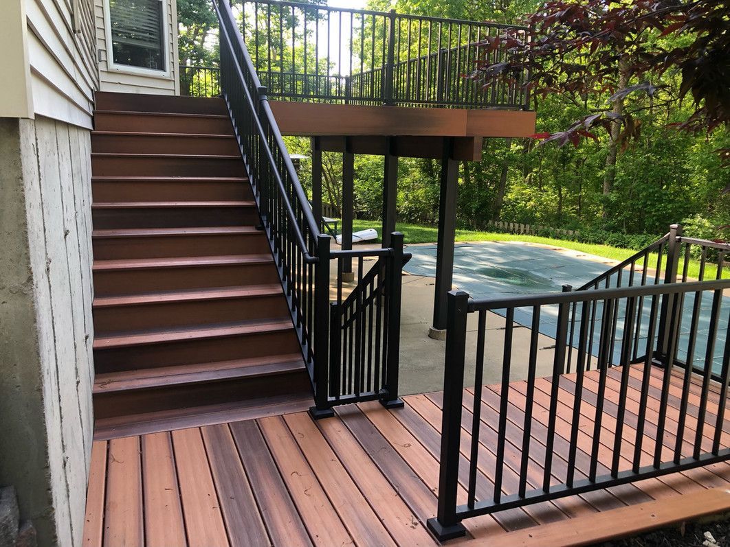 Wooden deck and stairs with black railing leading to a raised deck, surrounded by green trees.
