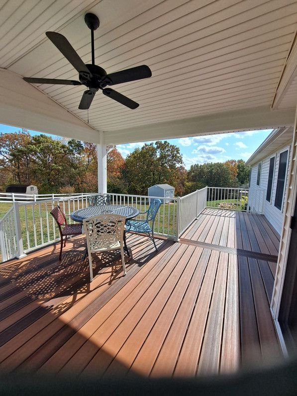 Covered outdoor deck with seating, ceiling fan, overlooking a yard with fall foliage.