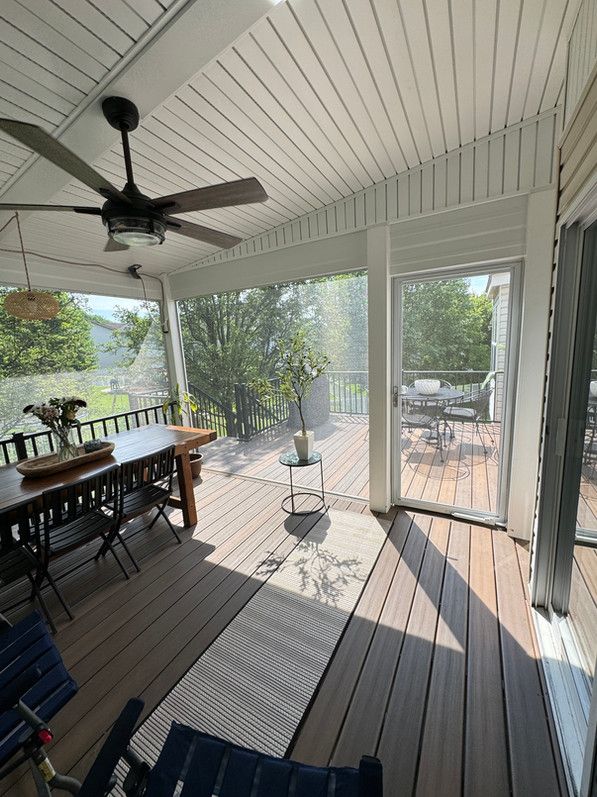 Screened porch with dining table, rug, ceiling fan, and view of trees and deck outside.