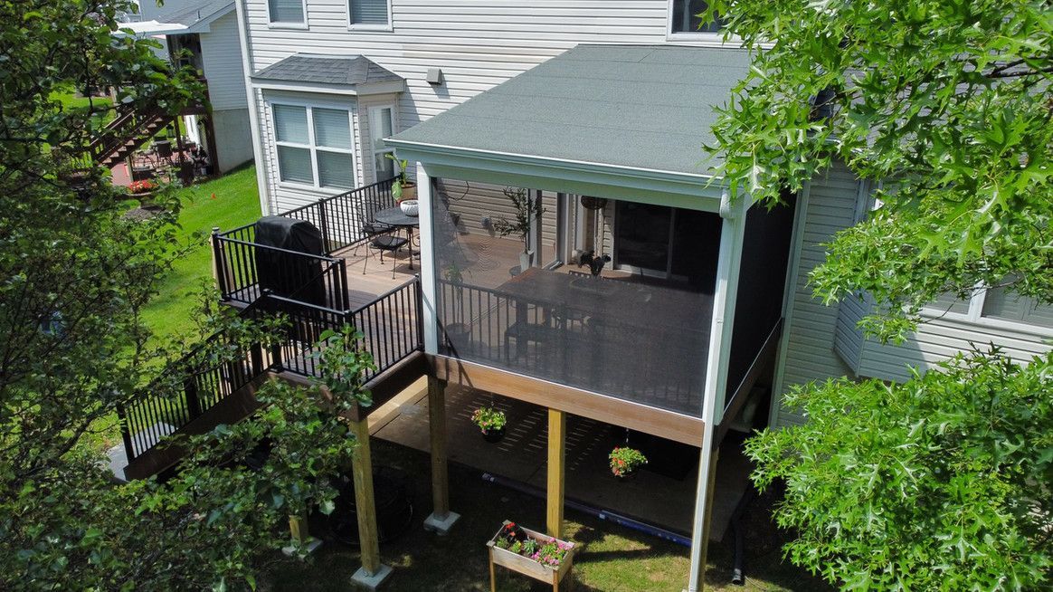 Screened porch with dark deck and attached grill next to a two-story house, surrounded by green trees and grass.