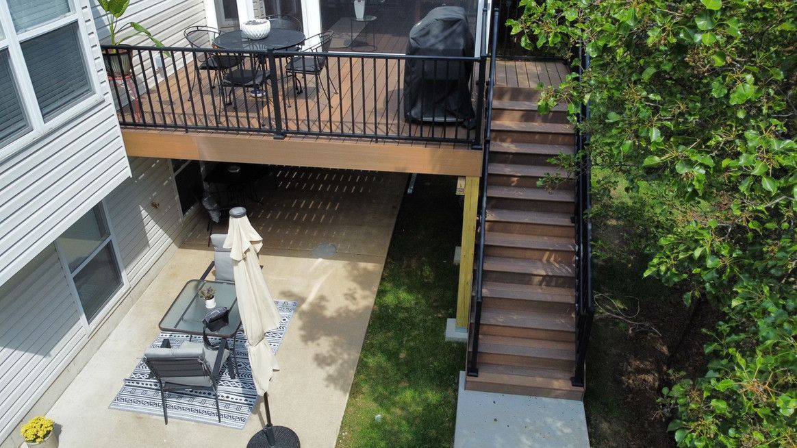 Exterior shot of a deck with stairs. A patio is below, and a grassy area with a tree is to the right.