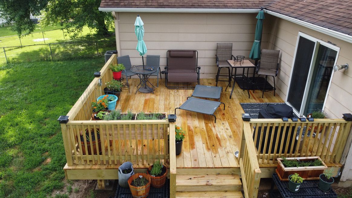 Wooden deck with outdoor furniture, including tables, chairs, and a chaise lounge, beside a house with a sliding door.