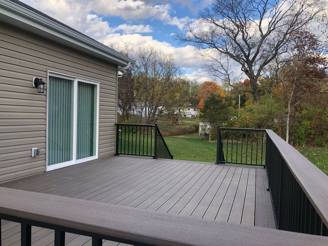 Gray composite deck with black railing and sliding glass door, in front of a green yard and trees.