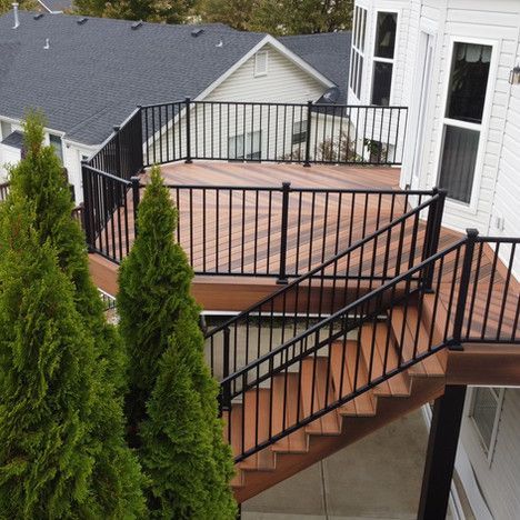 Wooden deck with black railings and stairs. Two green trees in the foreground. Houses in the background.