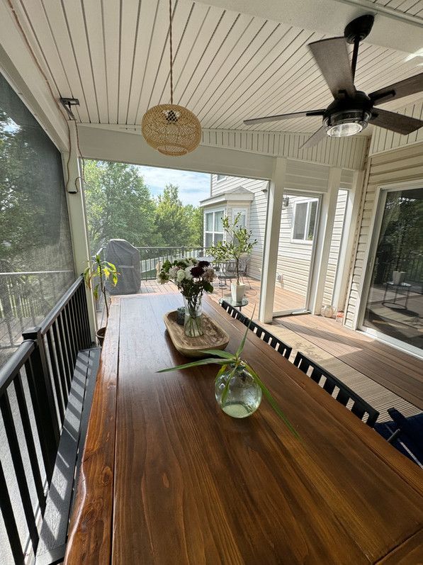 Screened-in porch with a dark wood table, flowers, pendant light, and ceiling fan. Overlooks a deck and wooded area.