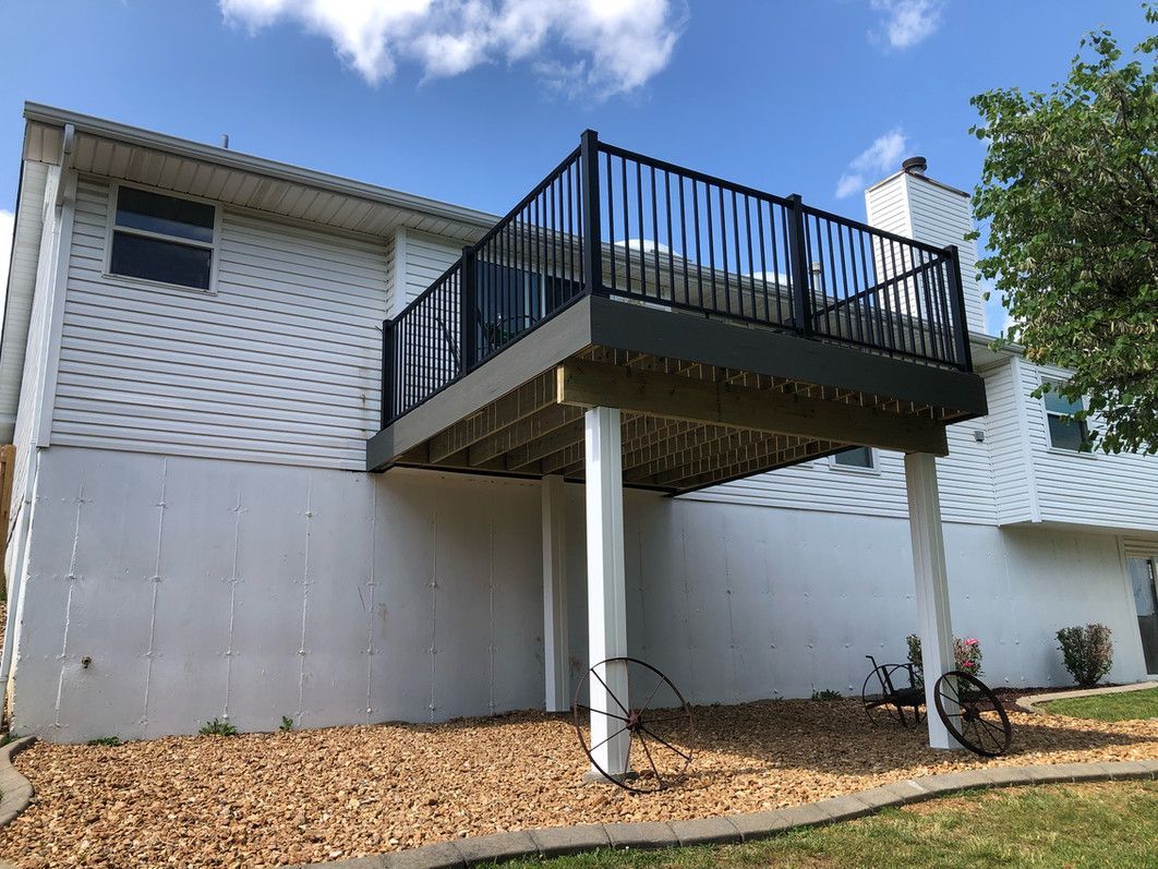 White house with elevated deck featuring black railing, supported by white posts, set on gravel.