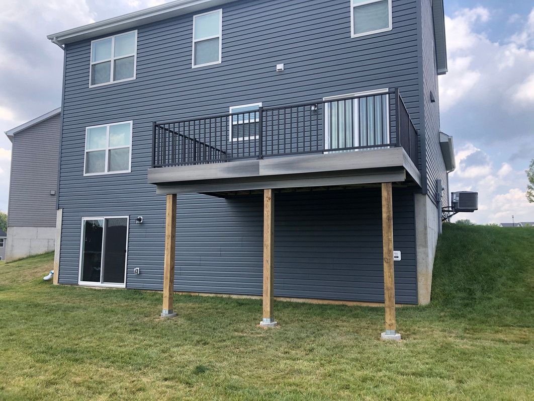 Two-story house with a gray deck and railing supported by wooden posts on a grassy lawn.
