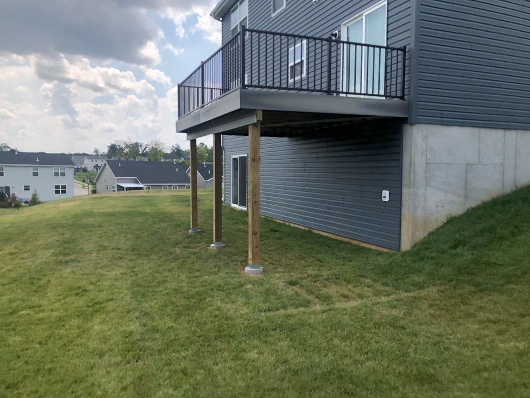 Back view of a house with a deck supported by wooden posts on a grassy hill under a cloudy sky.