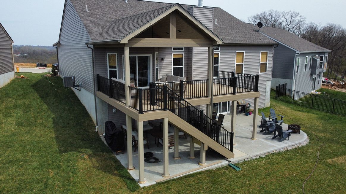 Two-story deck attached to a gray house, with a lower concrete patio, set on a grassy hill.