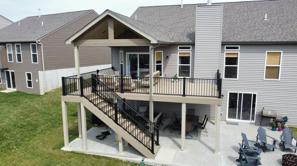 Two-story house with elevated deck and patio. Gray siding, black railing, brown deck. Green grass.