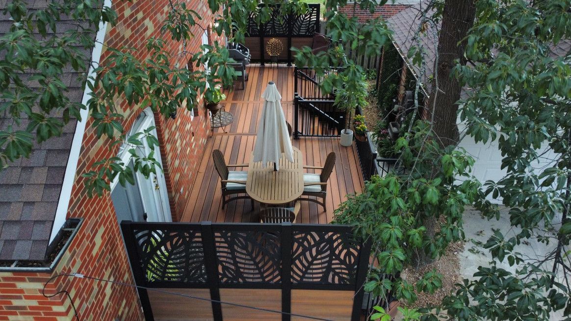 Wooden deck with furniture, umbrella, and decorative railing, surrounded by brick and trees.
